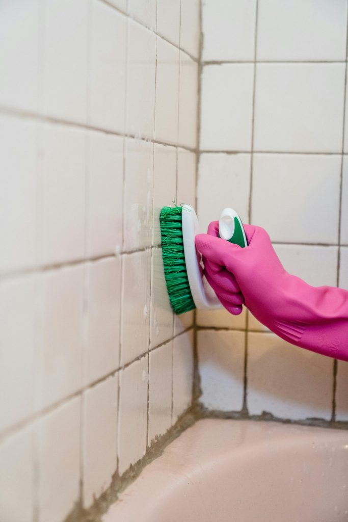 Eine Person schrubbt Badezimmerfliesen und trägt dabei einen rosa Handschuh, um die Sauberkeit zu betonen.